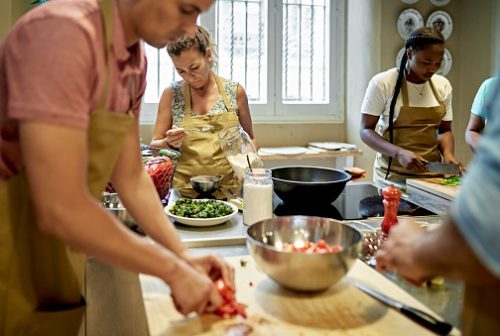 Focus on background businesswomen in aprons slicing and seasoning ingredients for dish prepared during team cooking class.